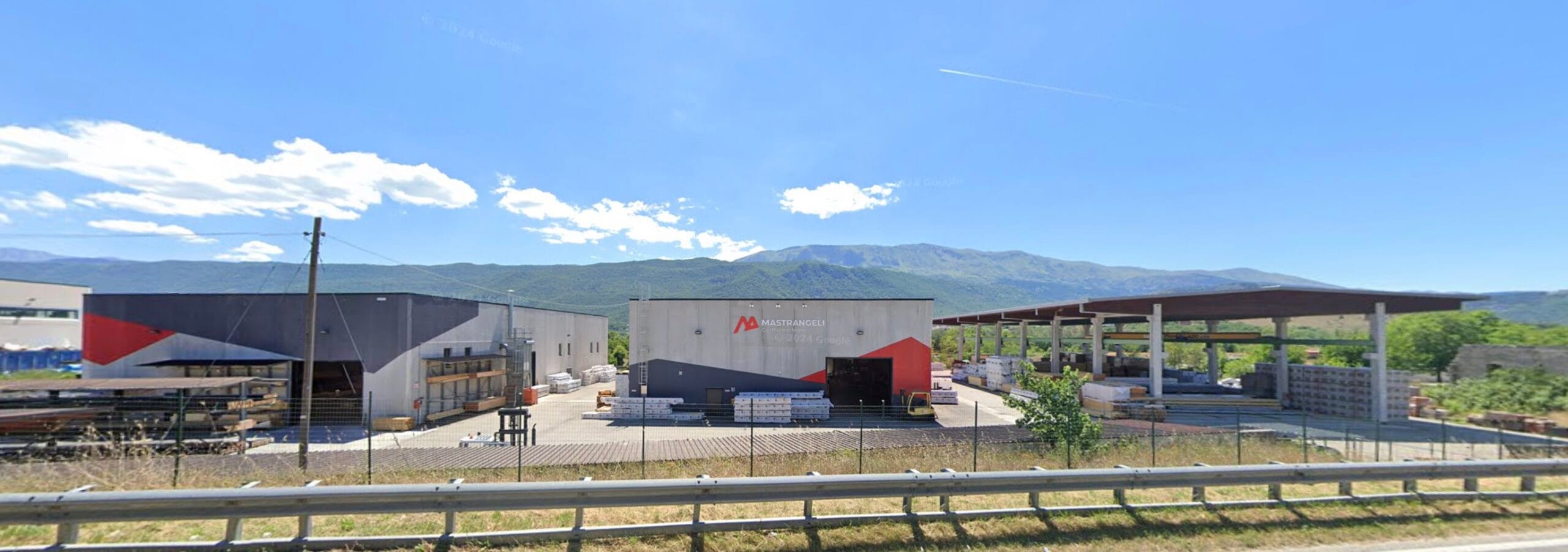 Capannone industriale in cemento con materiali da costruzione all’esterno, ai piedi di una catena montuosa sotto un cielo sereno.
