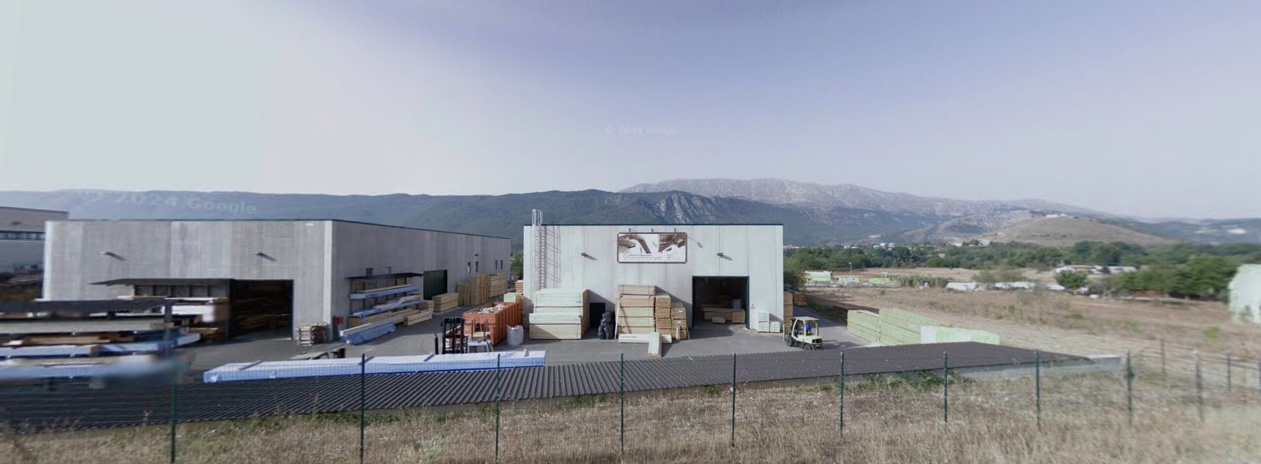 Capannone industriale in cemento con materiali da costruzione all’esterno, ai piedi di una catena montuosa sotto un cielo sereno.