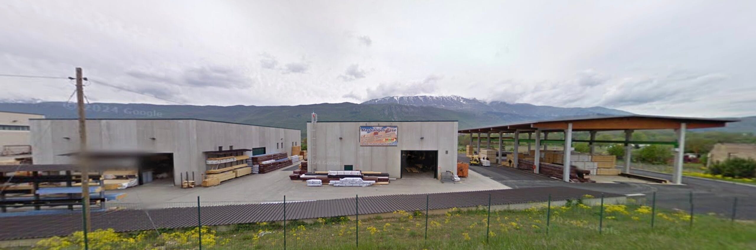 Capannone industriale in cemento con materiali da costruzione all’esterno, ai piedi di una catena montuosa sotto un cielo sereno.